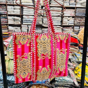 Może przedstawiać: Duża torba typu tote z żywym wzorem przedstawiającym powtarzający się wzór tygrysów w kolorze czarnym i złotym na różowo-czerwonym tle w paski. Torba ma czerwone paski i wykończenia i wisi na brązowym wieszaku.