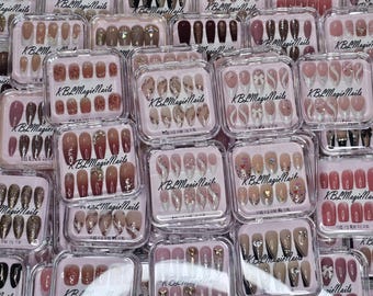 May include: An assortment of artificial nail sets in clear plastic cases. The sets feature various designs, including solid colors, glitter, and patterns. The text "KBL Magie Nails" is printed on each case.