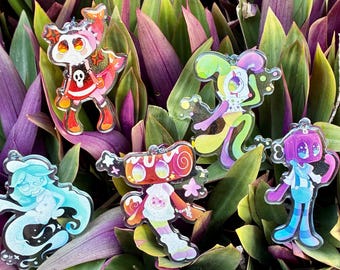 May include: Five clear acrylic charms featuring colorful cartoon characters. One has a skull design, one is a clown-like figure, one is a blue ghost-like form, one has a swirl pattern, and one is a purple-headed figure with blue striped legs.