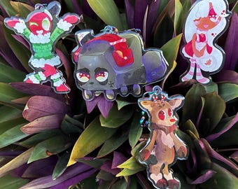 May include: Four acrylic keychains featuring cartoon characters: a red figure in a green sweater, a dark gray block-shaped creature with red headphones, a white-haired character in a red dress, and a brown reindeer with red antlers and nose.