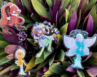 May include: Four acrylic charms featuring stylized character illustrations. One is a brown creature, one is a floral-themed figure, one is a small orange-headed character, and one is a white and teal moth-like being with glittery accents.
