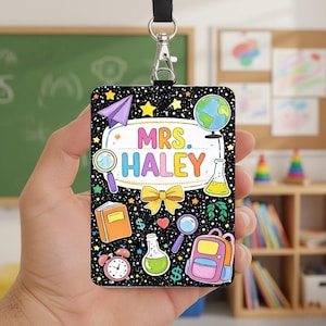Könnte beinhalten: Ein schwarzes Schlüsselband mit einem rechteckigen Abzeichen mit farbenfrohen Illustrationen zum Thema Schule. Das Abzeichen trägt den Text "MRS. HALEY" in einer Regenbogen-Schriftart, umgeben von Sternen, einem Papierflieger, einem Globus und anderen Schulmaterialien.