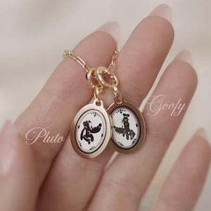 May include: A gold-colored necklace featuring two oval pendants. Each pendant has a black and white cartoon character design. The word "Pluto" is written on the left side, and "Goofy" is written on the right side.