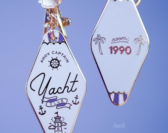 Könnte beinhalten: Zwei weiße und goldene Schlüsselanhänger in Diamantform. Einer trägt die Aufschrift "Ahoy Captain Yacht Club" mit Leuchtturm und Anker. Der andere trägt "room: 1990" mit Palmen. Beide haben goldene Ketten.