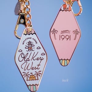 May include: Two diamond-shaped keyrings with gold-coloured chains and clasps. One keyring is white with "Old Key West" text and a lighthouse graphic. The other is pink with "room: 1991" text and palm tree graphics. The front and back are visible.