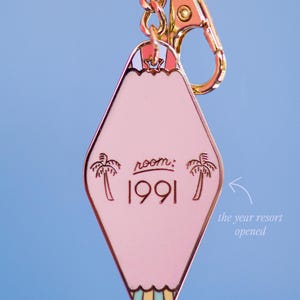 May include: Pink motel-style keychain with gold-tone hardware. The keychain has a diamond shape with the text "room: 1991" and palm tree graphics. The back of the keychain is visible.