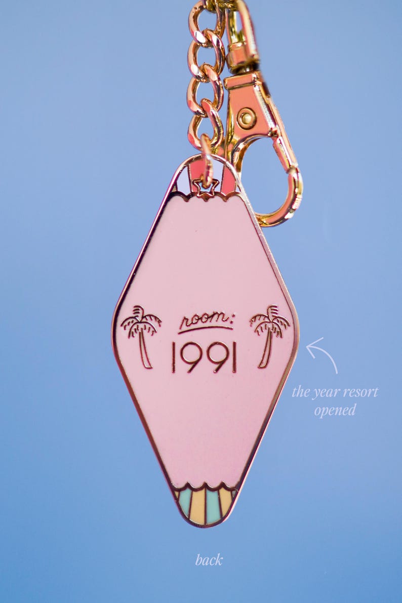 May include: Pink motel-style keychain with gold-tone hardware. The keychain has a diamond shape with the text "room: 1991" and palm tree graphics. The back of the keychain is visible.