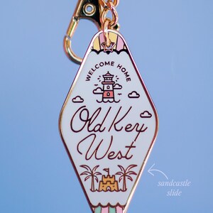 May include: A diamond-shaped keyring with a gold chain and clasp. The white enamel face features a lighthouse, clouds, and the words "WELCOME HOME" and "Old Key West". A sandcastle slide is at the bottom.