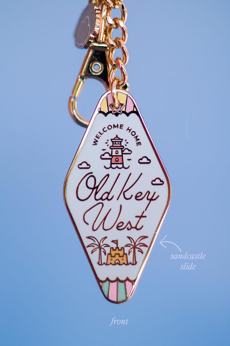May include: A diamond-shaped keyring with a gold chain and clasp. The white enamel face features a lighthouse, clouds, and the words "WELCOME HOME" and "Old Key West". A sandcastle slide is at the bottom.