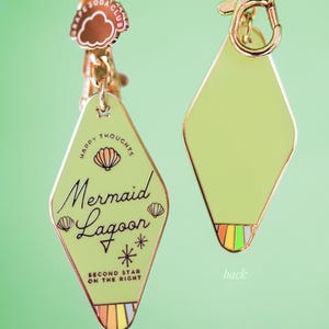 Puede incluir: Dos llaveros con una etiqueta en forma de diamante verde claro. La etiqueta frontal dice "Mermaid Lagoon" con gráficos de conchas marinas y estrellas. La etiqueta trasera es de color verde claro liso. Ambos tienen herrajes dorados y un dije que dice "Soda Club".