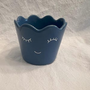 May include: A blue ceramic planter with a wavy rim and a smiling face design. The planter features white eyelashes and a curved mouth, adding a whimsical touch. This decorative pot is suitable for small plants or succulents.