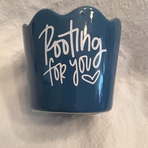 May include: A small, blue ceramic planter with a scalloped rim. White cursive text on the front reads "Rooting for you" with a heart symbol. The planter is designed for small plants or succulents.