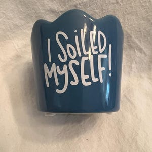 May include: A small, blue ceramic planter with a scalloped top edge. The white text "I SOILED MYSELF!" is printed on the front. The planter is designed for small plants or succulents.