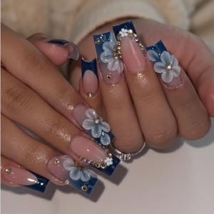 May include: Long square-shaped artificial nails featuring a dark navy blue French tip design on a nude base. The nails are decorated with three-dimensional light blue floral accents, small white flowers, and clear rhinestones embedded near the cuticles.