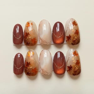 May include: A set of ten oval-shaped false nails. The nails feature a variety of designs, including solid brown, shimmering copper, and clear with gold leaf maple leaves. Some nails have gold wire accents. The overall color palette is warm, with shades of brown, copper, and gold.