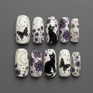 Pode incluir: Um conjunto de dez unhas postiças com base creme e ilustrações em preto e roxo. Os designs incluem gatos pretos, borboletas, luas crescentes e padrões florais roxos com pequenos detalhes de estrelas, dispostos em duas filas de cinco.