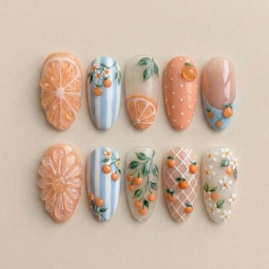 May include: A collection of ten almond-shaped acrylic nails with various designs. The designs include orange slices, orange blossoms, and orange-themed patterns. Colors include orange, peach, white, and blue. Some nails feature 3D elements.