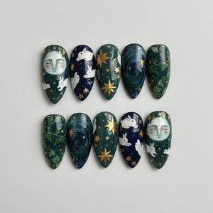 May include: A set of ten almond-shaped press-on nails featuring hand-painted designs. The artwork includes white rabbits, golden stars, a moon face with closed eyes, swirling dark blue nebula patterns, and green ivy leaves on dark backgrounds.