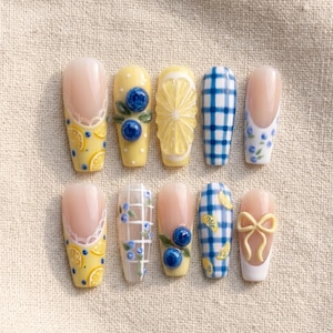 Puede incluir: Un juego de diez uñas postizas con varios diseños, incluyendo bases amarillas con rodajas de limón, patrones de cuadros vichy azules y blancos, detalles de arándanos en 3D, encaje blanco y un lazo 3D amarillo pálido sobre base nude.