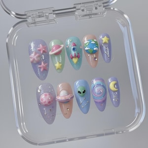 May include: A set of ten artificial nails in a clear plastic case. Each nail features a different space-themed design, including planets, stars, rockets, and an alien. The nails are painted in pastel colors with glitter accents.