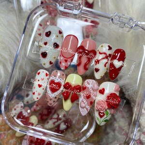 May include: A set of ten almond-shaped press-on nails featuring various designs including red and pink hearts, white lace patterns, red bows, and a three-dimensional red rose. The nails are displayed in a clear plastic container with MaydayStudioUs text.