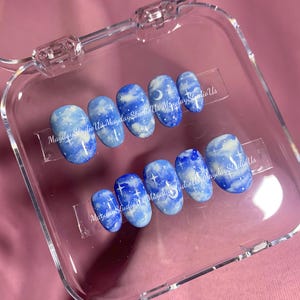 May include: A set of ten press-on nails featuring a blue sky design with white clouds, crescent moons, and four-pointed stars. The nails are arranged in two rows inside a clear plastic case, with the text MaydayStudioUs printed across them.