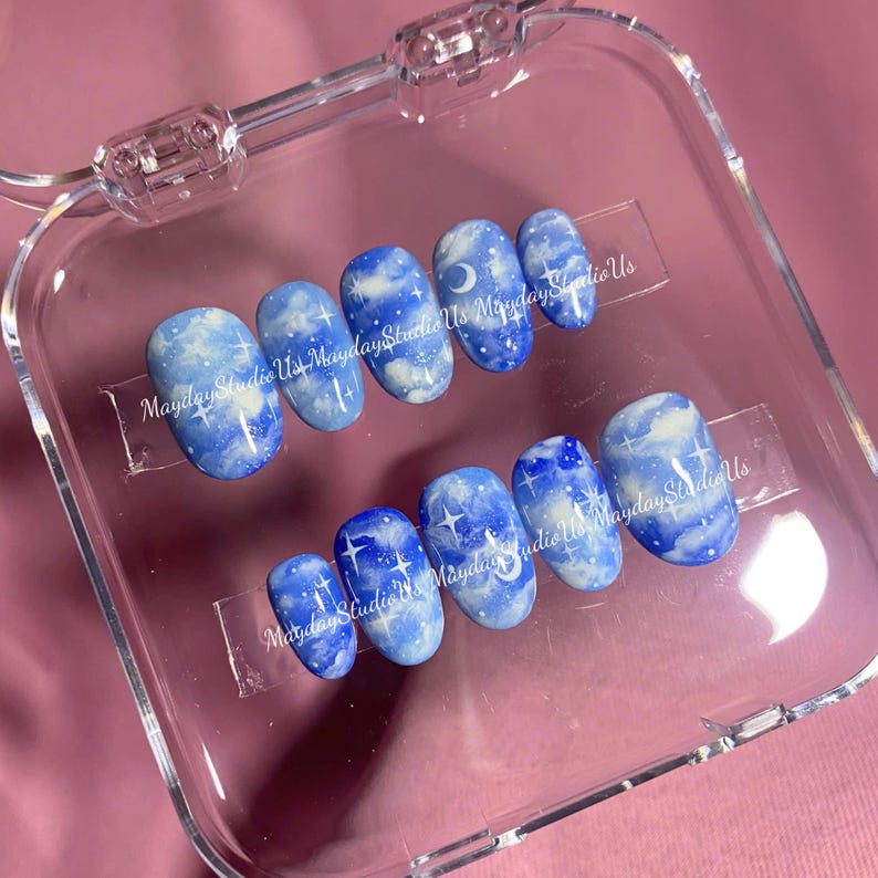 May include: A set of ten press-on nails featuring a blue sky design with white clouds, crescent moons, and four-pointed stars. The nails are arranged in two rows inside a clear plastic case, with the text MaydayStudioUs printed across them.