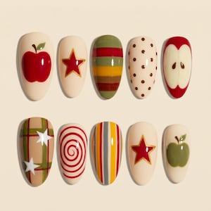 Puede incluir: Un conjunto de diez uñas postizas en forma de almendra con varios diseños, incluyendo una manzana roja, una estrella roja, rayas horizontales, puntos marrones, una rodaja de manzana, un patrón de cuadros verde con estrellas blancas y una espiral roja.