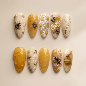 Puede incluir: Un conjunto de diez uñas artificiales en forma de almendra con un tema de abeja y miel. Los diseños incluyen abejas, margaritas, panales y miel goteando, en tonos de amarillo, blanco y dorado. Las uñas están dispuestas sobre una superficie beige claro.