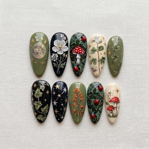 May include: A set of ten almond-shaped press-on nails featuring intricate hand-painted designs including a full moon, white flowers, red-spotted mushrooms, green ivy vines, gold stars, constellations, and small red ladybugs on olive, black, and cream bases.
