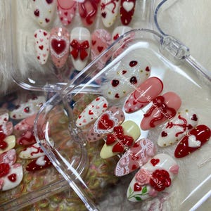 May include: A set of almond-shaped press-on nails featuring a Valentine's theme with red and pink hearts, 3D red bows, white lace patterns, and floral accents. The nails are displayed in a clear plastic container with the text MaydayStudioUs.