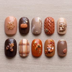 May include: A set of ten press-on nails featuring autumn-themed designs including a gold leaf, a 3D pumpkin, marble patterns, a cable knit texture, a squirrel, acorns, plaid, dried flowers with gold foil, a fox, and a glitter-bordered leaf.