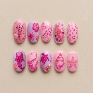May include: A set of ten oval press-on nails featuring various pink and lavender designs. The patterns include sharks, hibiscus flowers, sea turtles, seashells, starfish, and bubbles, with some nails accented by small pearls and textured details.