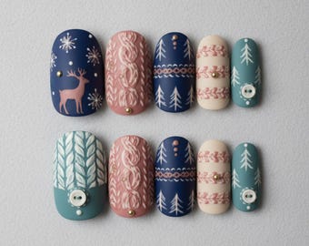 May include: A set of ten artificial nails with winter-themed designs. The nails feature patterns like snowflakes, a reindeer, cable knit, and Christmas trees in shades of blue, pink, and cream. Some nails have small gold accents and button details.