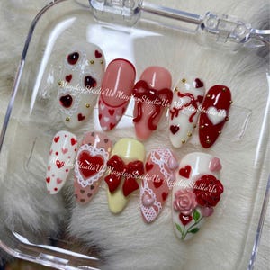 May include: A set of ten press-on nails featuring various designs including red hearts, white lace patterns, 3D red bows, and a 3D red rose. The nails are arranged in a clear plastic case, each decorated with small gold studs and heart motifs.