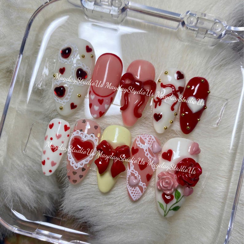 May include: A set of ten press-on nails featuring various designs including red hearts, white lace patterns, 3D red bows, and a 3D red rose. The nails are arranged in a clear plastic case, each decorated with small gold studs and heart motifs.