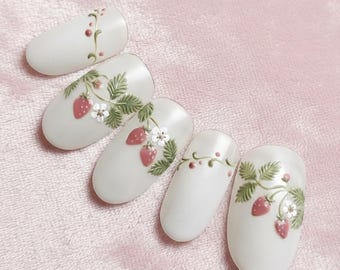 Könnte beinhalten: Fünf weiße Kunstnägel mit Erdbeerdesign. Jeder Nagel hat ein anderes Erdbeer- und Blättermuster in Rot, Grün und Weiß. Die Nägel sind auf einer rosa Oberfläche angeordnet.