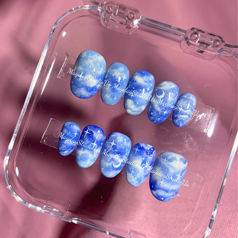May include: A set of ten press-on nails in a clear plastic case, each featuring a blue sky design with white clouds, stars, and crescent moons. The text MaydayStudioUs is printed across the nails in a white script font, showcasing a celestial theme.