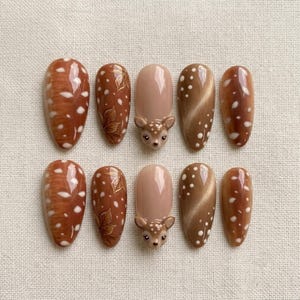 Op de afbeelding: Een set van tien kunstnagels in bruin- en beigetinten. De ontwerpen bevatten effen kleuren, witte stippen, geschilderde bladpatronen en een driedimensionaal gebeeldhouwd hertenkopje op de middelste nagel met een glanzende afwerking.