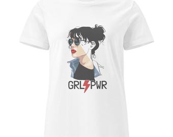 Può includere: T-shirt bianca con grafica di una donna con occhiali da sole e giacca di jeans. Il testo "GRL PWR" è sotto l'illustrazione con un fulmine rosso. La maglietta ha maniche corte e girocollo.