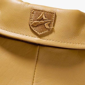 以下が含まれることがあります： 茶色の革製品のクローズアップ。ステッチされたエンブレムが付いています。エンブレムは盾の形で、詳細な刺繍デザインが施されています。革は微妙な質感です。