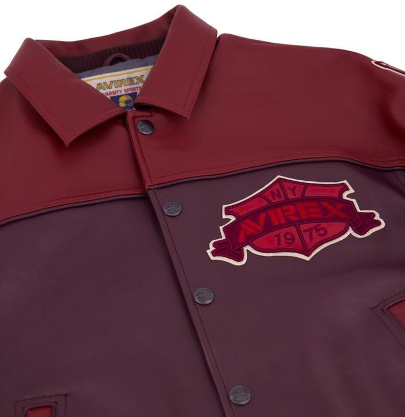 Może przedstawiać: Bordowa i kasztanowa kurtka w stylu college z zapięciem na guziki i kołnierzem. Kurtka posiada naszywkę z napisem "NY AVIREX 1975" w kształcie tarczy. Kurtka wykonana jest z materiału przypominającego sk&oacute;rę.