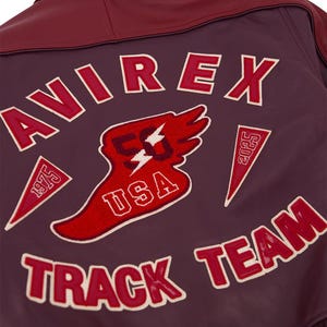 Może przedstawiać: Zbliżenie na tył bordowej sk&oacute;rzanej kurtki z napisem "AVIREX" w czerwonych literach z białymi obw&oacute;dkami. Kurtka posiada kilka czerwono-białych naszywek, w tym "USA", "TRACK TEAM" i "1975".