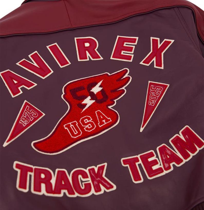 Może przedstawiać: Zbliżenie na tył bordowej sk&oacute;rzanej kurtki z napisem "AVIREX" w czerwonych literach z białymi obw&oacute;dkami. Kurtka posiada kilka czerwono-białych naszywek, w tym "USA", "TRACK TEAM" i "1975".