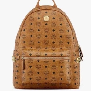 Puede incluir: Mochila marrón con un patrón repetido de las letras "MCM" y formas de diamante. La mochila tiene un asa superior, un bolsillo frontal con cremallera y herrajes dorados. Es un diseño clásico.
