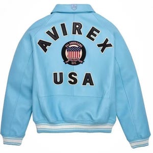 Puede incluir: Chaqueta azul claro con la palabra "AVIREX" en letras negras en la espalda. Debajo, un parche con "ALL AMERICANS" y "USA" en negro. La chaqueta tiene pu&ntilde;os a rayas blancas y azules y un cuello.
