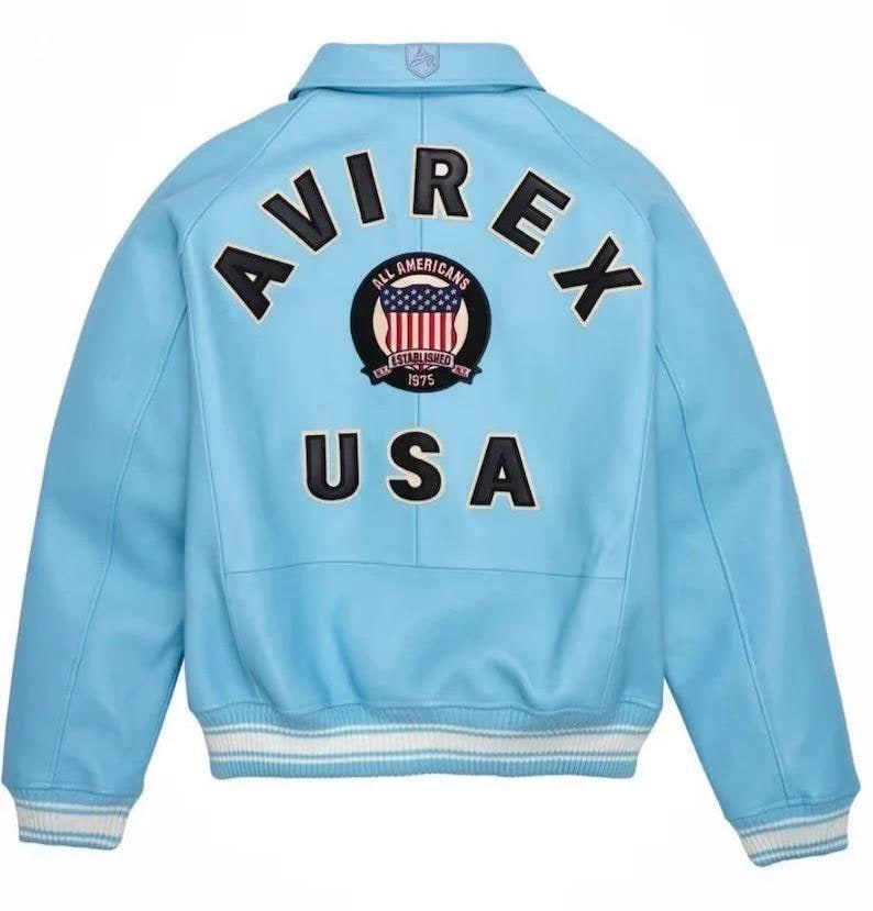 Puede incluir: Chaqueta azul claro con la palabra "AVIREX" en letras negras en la espalda. Debajo, un parche con "ALL AMERICANS" y "USA" en negro. La chaqueta tiene pu&ntilde;os a rayas blancas y azules y un cuello.