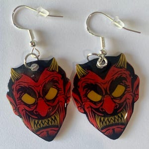 May include: A pair of devil-themed earrings. Each earring features a red devil face with yellow eyes and teeth, black hair, and yellow horns. The earrings hang from silver-colored fish hook ear wires.