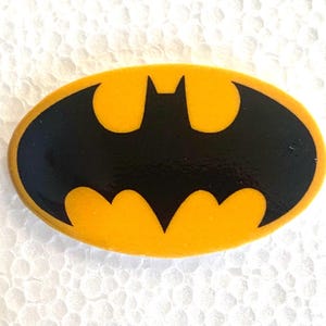 May include: Oval Batman logo badge. The badge displays the black bat symbol on a yellow background. The design is a classic representation of the DC Comics superhero.