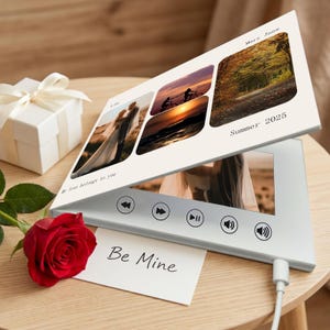 May include: A white digital photo frame showing several images, with the text "Mary Jane" and "Summer 2025". A red rose and a "Be Mine" card are in the foreground. A white gift box with a bow is also present.